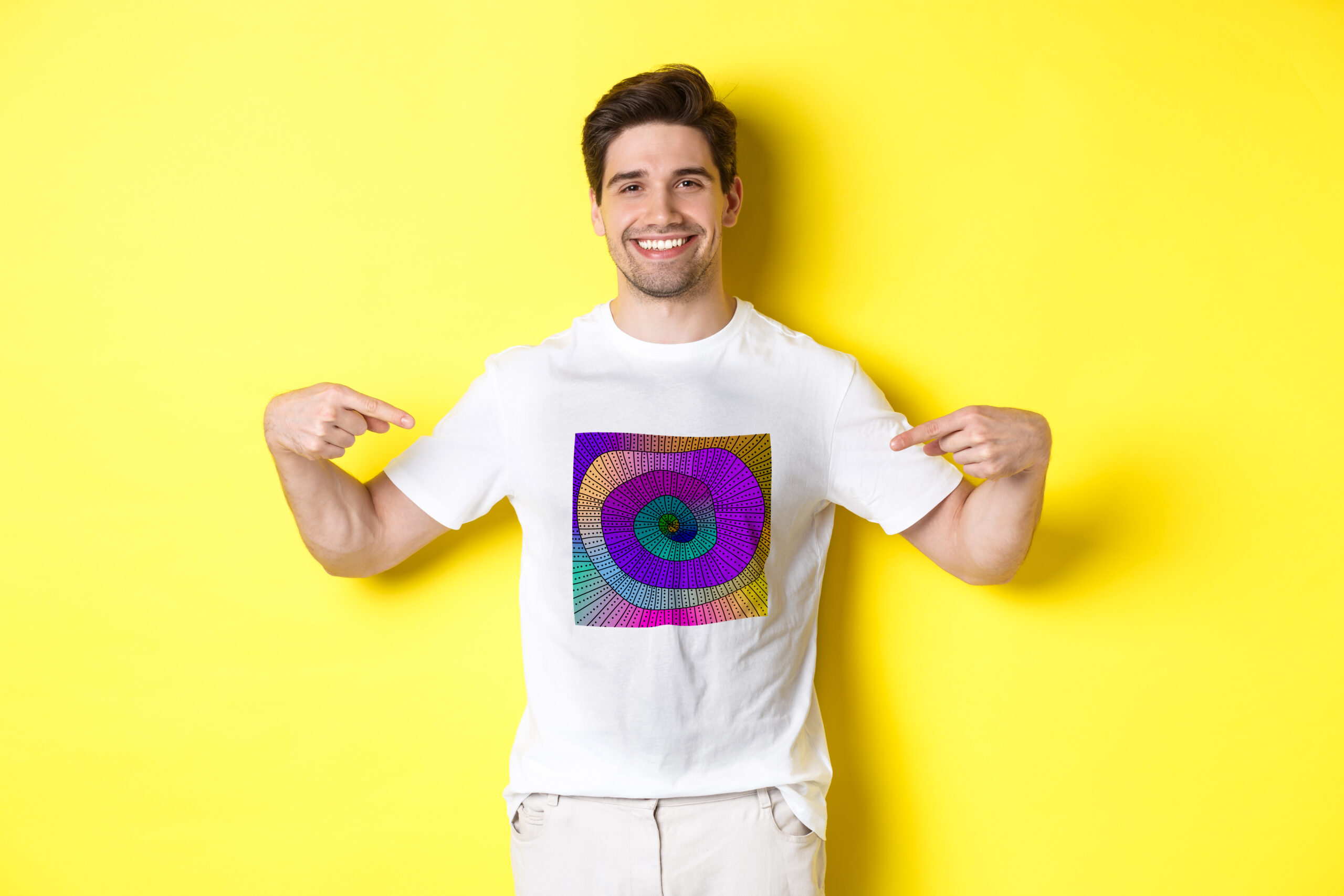 Happy attractive guy pointing fingers at your logo, showing promo on his t-shirt, standing over yellow background.